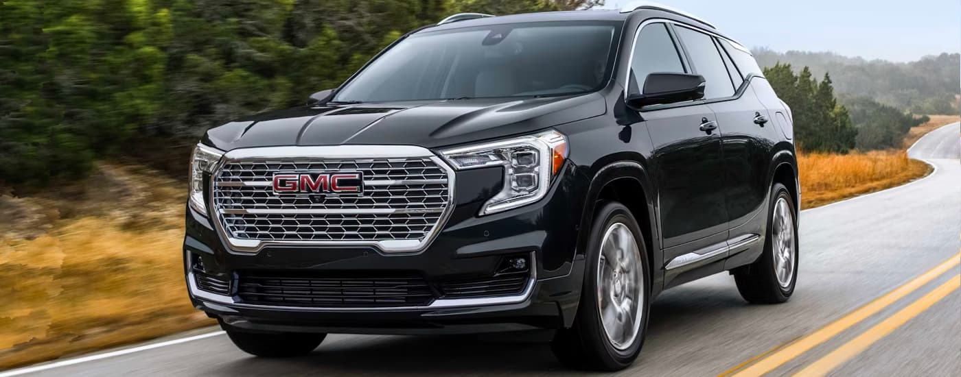 Black 2023 GMC Terrain Denali driving from a used car dealership.